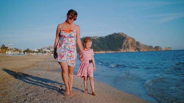 Mother wearing colorful dress with her happy daughter holding hands walking on beach along coastline. Clear blue sky, mountains and sea waves in background. Slow motion.