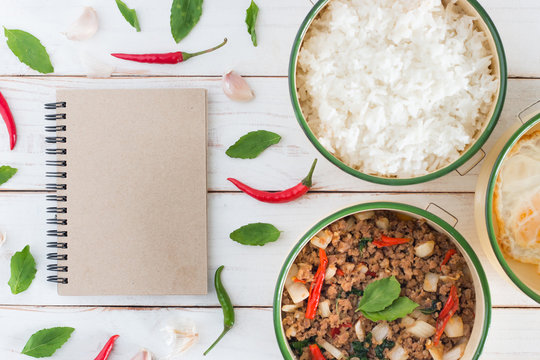 Thai food name Pad Ka Prao,Top view image of Stir-fried pork with basil leaves beside have Cooked rice and Fried egg in classic food carrier with brown notebook set on white wood table background