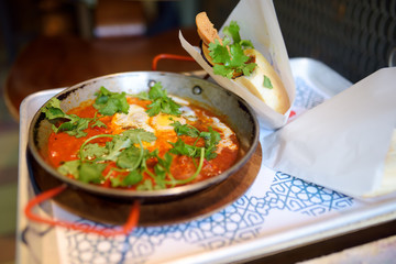 Shakshuka at pan on tray in cafe of Israeli cuisine. Shakshouka is fried eggs with tomatoes, vegetables and herbs.