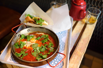 Shakshuka at pan on tray in cafe of Israeli cuisine. Shakshouka is fried eggs with tomatoes, vegetables and herbs.