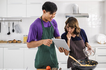 Smiling happy young Asian couple wearing apron and cooking together in a white kitchen background.