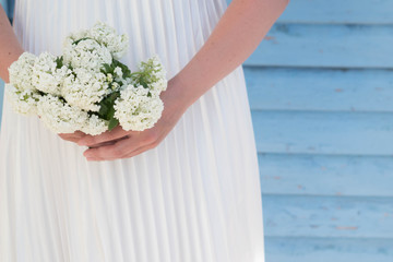 Slender woman in white satin pleated dress holding a small bouquet of white flowers in hands on a blue lath background with copyspace. Use for card design, for flower shop, cosmetic, wedding store