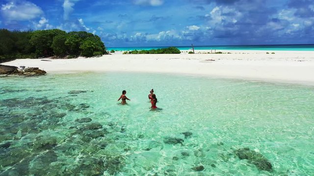 Clear turquoise water with rocks in the foreground, in which three young women in red swimsuits enjoying the Caribbean sea. A sandbank in the background and a dramatic Sky. Aerial dolly in, wide angle