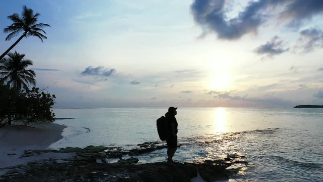 silhouette of young tourist with backpack admiring majestic sunrise on tropical beach with palms,dolly in full shot