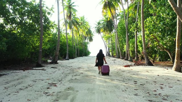 dolly in shot of young woman with her luggage just arriving on tropical Caribbean island. Walking on sandy path thru coconut palm forest