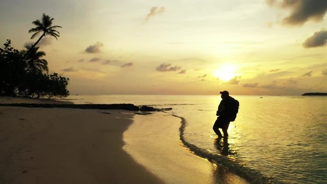 A silhouette of a man wearing a backpack and a cap is staying in shallow water while looking onto a Caribbean island. Iit by the golden light of the setting sun. Wide angle, dolly out.