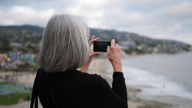 A middle aged woman on vacation taking a picture with her phone of the city and ocean in Laguna Beach, California SLOW MOTION.