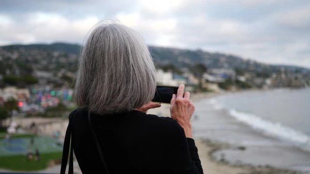 An elderly woman on vacation taking a picture with her phone of the city and ocean in Laguna Beach, California SLOW MOTION.