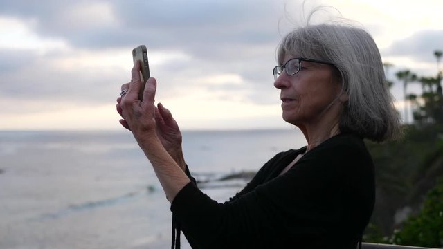 An old lady on vacation happy and smiling while taking a selfie with her smart phone on the ocean in Laguna Beach, California SLOW MOTION.