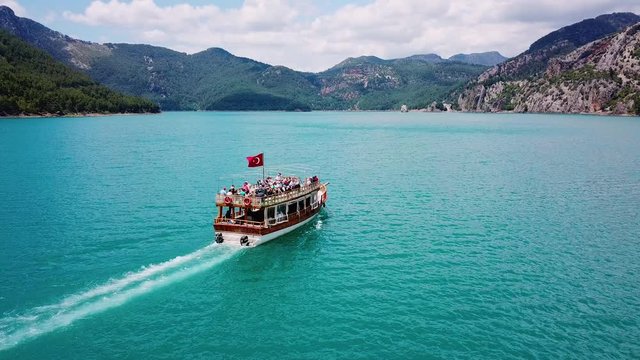 Aerial view: sightseeing boat with tourists sailing on the reservoir Green Canyon Antalya Turkey with clear turquoise water surrounded by mountains and forest.