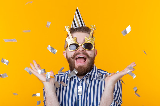 Joyful young hipster male in a paper cap and glasses is laughing happily among flying confetti on a yellow background. The concept of a mega party and holiday.