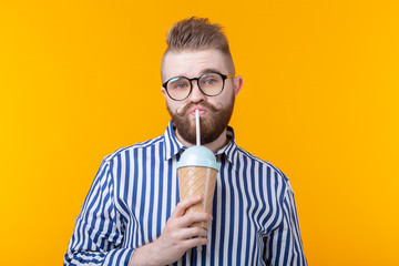 Cute funny young man with a mustache and beard drinking a cocktail with a straw on a yellow background. The concept of summer desserts and drinks.