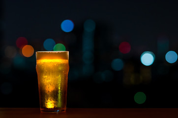 A glass of Beer with its foam puts on wooden table of the bar isolated on dark night background with colorful bokeh lights on rooftop bar.