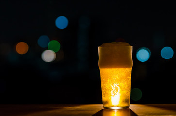 A glass of Beer with its foam puts on wooden table of the bar isolated on dark night background with colorful bokeh lights on rooftop bar.