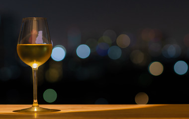 A glass of white wine on wooden table of rooftop bar with colorful bokeh of city light and space for text.