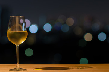 A glass of white wine and its shadow on wooden table of rooftop bar with colorful bokeh of city light and space for text.