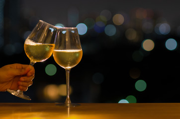 Hand holding a glass of white wine toasting to celebration and party concept put on wooden table of rooftop bar with colorful bokeh of city light and space for text.