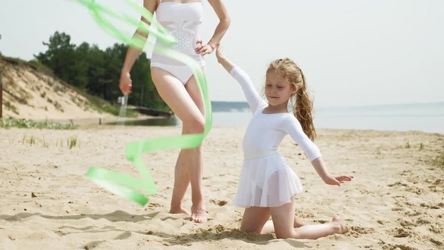 mother and daughter in white bathing suits dancing with gymnastic ribbon on a sandy beach. Summer, dawn