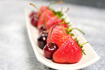 fresh sweet cherries and strawberries on a white plate. healthy food, vitamins. on the grey table, selective focus.