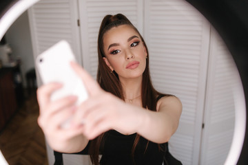 Young attractive model make a selfie after makeup in beauty studio. She use a ring lapm for photo. Background of white folding screen