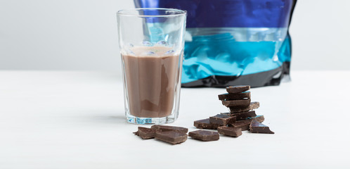 Glass mug with a protein chocolate shake stands against the background of a packet of protein on a white table. Sports nutrition concept.