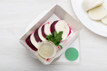 Top view of boiled beets with slices of white cheese lie in a white lunch box with sour cream sauce and parsley on a white table next to goat cheese. Protein snack concept.