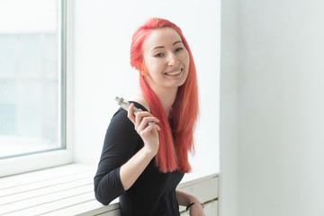 Youth and addiction concept - young red haired woman smoking vape near the window