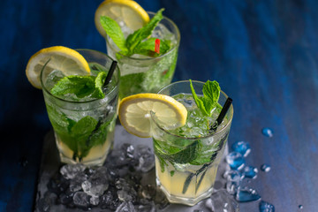 three glasses of mojito cocktail with fresh lemon and peppermint on a black stone board with ice