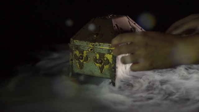 This slow motion video shows anonymous hands opening a mysterious treasure chest / pandoras box, revealing glowing mist / fog / smoke inside on black background