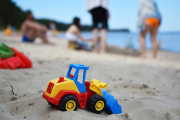 Toy tractor bulldozer on beach sand and blurred silhouette of people and sea on background. Leisure time with friends and family and summertime vacation concept.