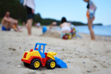 Fototapeta na wymiar Toy tractor bulldozer on beach sand and blurred silhouette of people and sea on background. Leisure time with friends and family and summertime vacation concept.