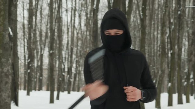 A handsome young man in a black suit and balaclava demonstrates mastery with nunchucks in an outdoor winter park. Goes in for sports and has fun