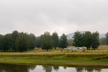 Heavy clouds in the cold autumn sky over  village with small light wooden houses far away in the mountains. Wide river flows near with reflections in the water. Travelling on the suburb roads. Fog