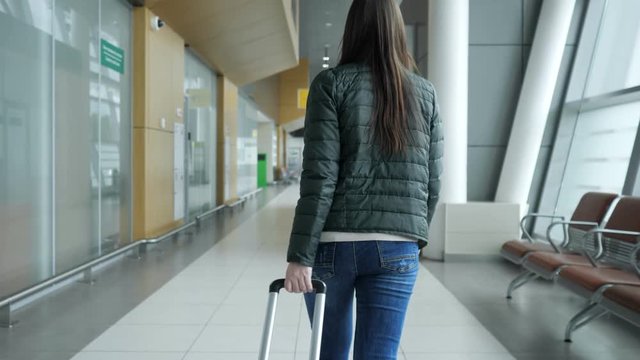 Traveler woman brunette is walking in modern airport terminal carrying suitcase, back view. Passenger is going to her flight in city airport.