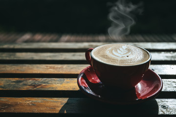 A cup of hot coffee or cappuccino is placed on an outdoor wooden table in the morning by the smoke rising from the red cup.