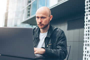 Fototapeta premium Attractive adult successful bald bearded man in black jacket with laptop in street cafe at city