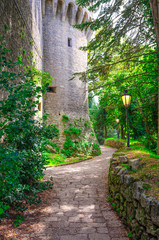 Cobblestone path with street light lamp near wall of stone brick medieval castle tower in green park forest in Republic San Marino