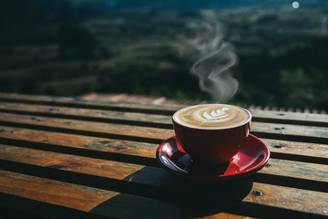 A cup of hot coffee or cappuccino is placed on an outdoor wooden table in the morning by the smoke rising from the red cup.