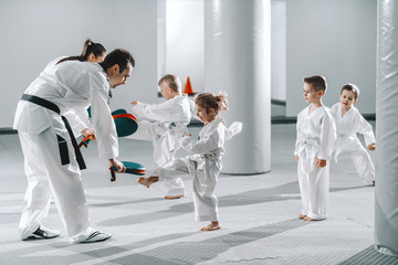 Small group of kids in doboks practicing with their trainers taekwondo moves while kicking in kick target.
