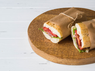 Fresh baguette sandwich with meat, sliced cheese, tomatoes and fresh lettuce in wrapping paper on white wood background. copy space