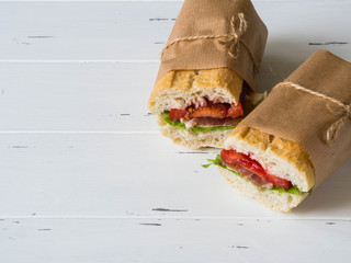 Fresh baguette sandwich with meat, sliced cheese, tomatoes and fresh lettuce in wrapping paper on white wood background. copy space