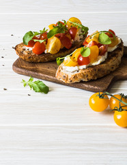 Pair of grilled toasts with cream cheese and slices of fresh tomatoes of various colors with fresh arugula and ground black pepper on a wooden cutting board
