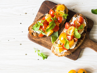 Pair of grilled toasts with cream cheese and slices of fresh tomatoes of various colors with fresh arugula and ground black pepper on a wooden cutting board