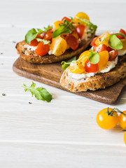Pair of grilled toasts with cream cheese and slices of fresh tomatoes of various colors with fresh arugula and ground black pepper on a wooden cutting board