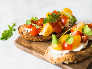 Pair of grilled toasts with cream cheese and slices of fresh tomatoes of various colors with fresh arugula and ground black pepper on a wooden cutting board