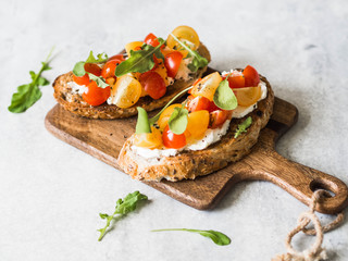 Pair of grilled toasts with cream cheese and slices of fresh tomatoes of various colors with fresh arugula and ground black pepper on a wooden cutting board