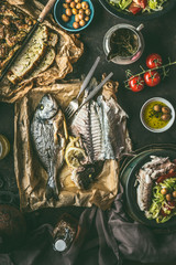Dinner table with backed dorado fish, salad and homemade bread on dark rustic table, top view. Mediterranean lunch or dinner. Healthy food