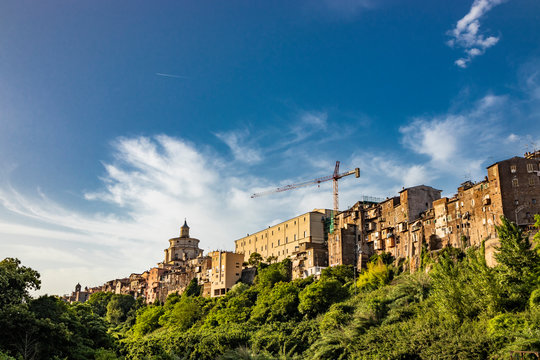 A view of the city of Zagarolo, with the houses built sheer above a tuff hill. Above the roofs, the dome of the church of San Pietro and a crane emerge. The valley full of trees. Rome, Lazio