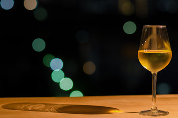 A glass of white wine and its shadow on wooden table of rooftop bar with colorful bokeh of city light and space for text.