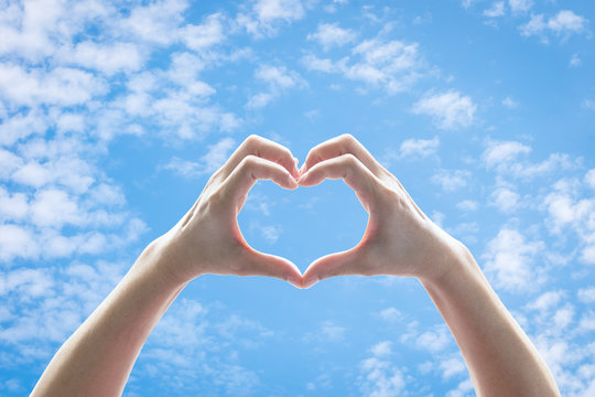 Woman hands in heart shape raising against blue sky with clouds for world kindness day, charity donation, friendship and Valentines love concept